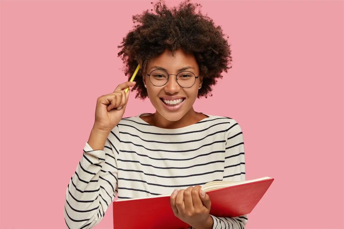 Bild eines schönen, fröhlichen Mädchens, das mit einem die Zähne zeigenden Lächeln Notizen mit einem Bleistift in ein Notizbuch schreibt.